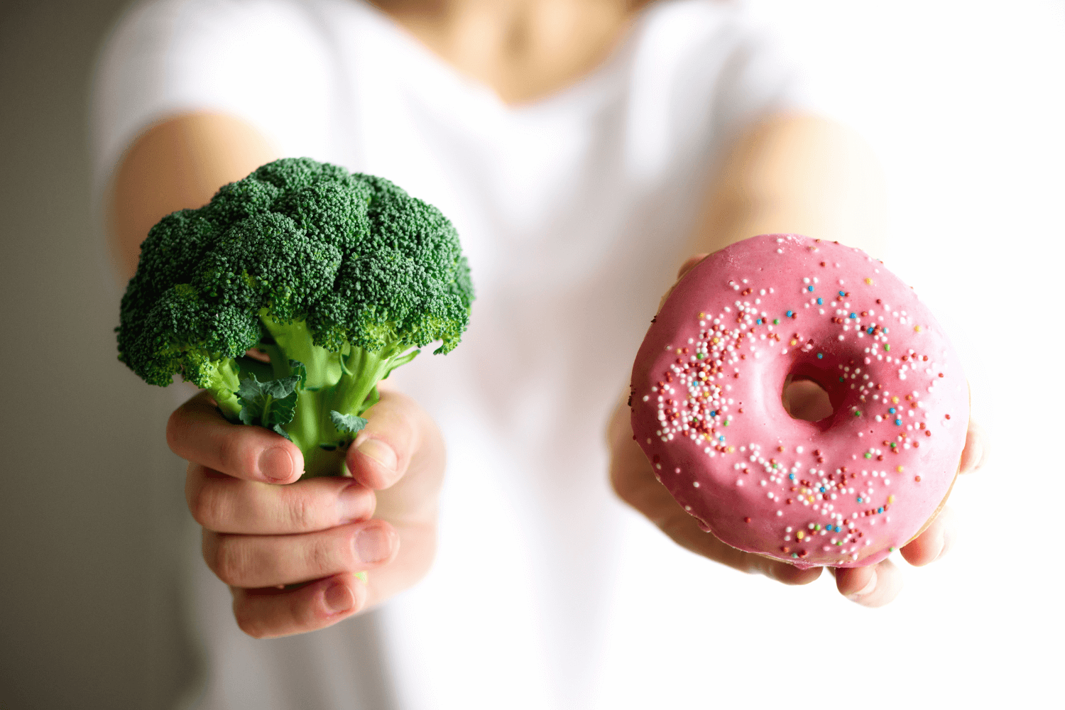 A person holds a stalk of broccoli in one hand and a donut in another.